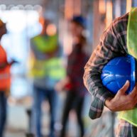 gettyimages-1178982949-612×612 Man holding blue helmet close up. Construction man worker with office and people in background. Close up of a construction worker's hand holding working helmet.