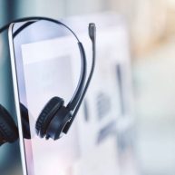 istockphoto-1400847561-612×612 Closeup of headset hanging on a computer desktop monitor in an empty call centre office. Operating helpdesk for customer service and sales support. Telemarketing agents providing online assistance